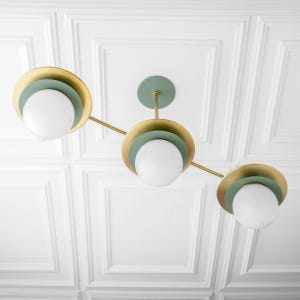 May include: A modern ceiling light fixture with three spherical white bulbs. The fixture features gold-colored circular accents with a light green interior, connected by thin gold rods. The fixture is mounted on a light green ceiling plate.