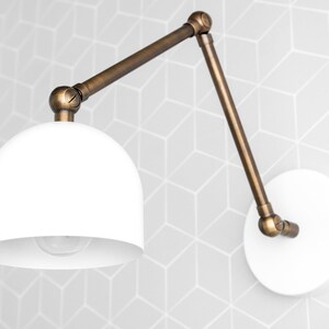 May include: A white, dome-shaped wall sconce with an adjustable, bronze-colored arm. The sconce is mounted on a white circular base. The background features a geometric pattern in shades of gray.