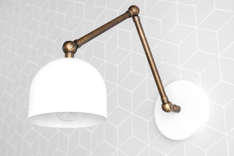 May include: A white, dome-shaped wall sconce with an adjustable, bronze-colored arm. The sconce is mounted on a white circular base. The background features a geometric pattern in shades of gray.