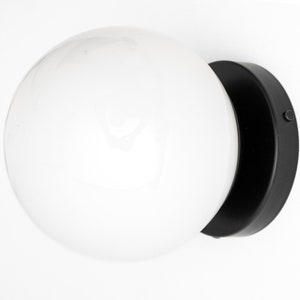 Peut inclure: Une applique murale noire avec un abat-jour en verre blanc en forme de globe. L'applique a une base ronde et un petit interrupteur sur le c&ocirc;t&eacute;.