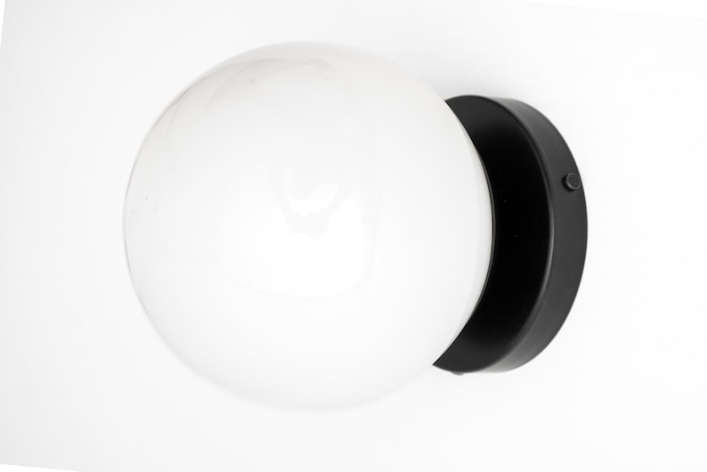 Peut inclure: Une applique murale noire avec un abat-jour en verre blanc en forme de globe. L'applique a une base ronde et un petit interrupteur sur le c&ocirc;t&eacute;.