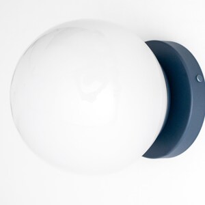 Peut inclure: Un luminaire globe blanc avec une base en m&eacute;tal bleu fonc&eacute;. Le luminaire est mont&eacute; sur un mur blanc.