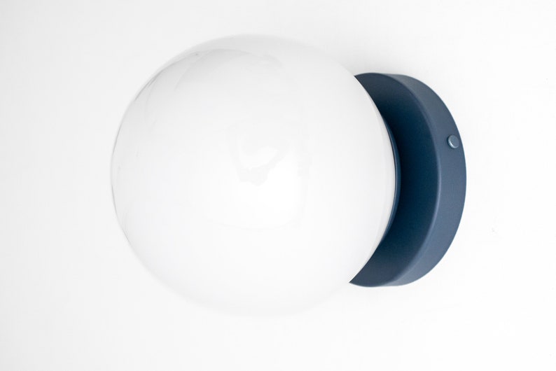 Peut inclure: Un luminaire globe blanc avec une base en m&eacute;tal bleu fonc&eacute;. Le luminaire est mont&eacute; sur un mur blanc.