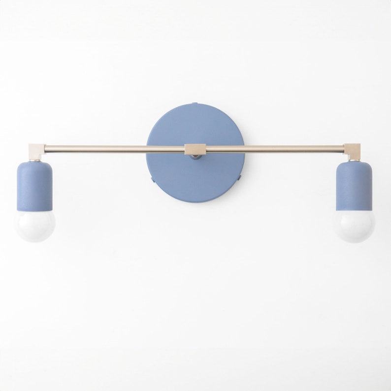 Puede incluir: Una l&aacute;mpara de pared azul y dorada con dos bombillas. La l&aacute;mpara tiene una placa trasera redonda azul y un brazo largo y recto con dos luminarias.