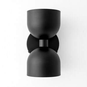 May include: Black modern wall sconce with a unique hourglass shape. The light fixture has two rounded, bowl-shaped shades connected by a short cylindrical piece. The sconce is mounted on a circular base.