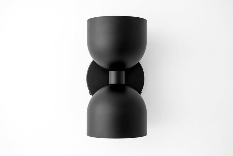 May include: Black modern wall sconce with a unique hourglass shape. The light fixture has two rounded, bowl-shaped shades connected by a short cylindrical piece. The sconce is mounted on a circular base.