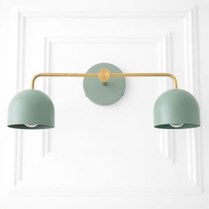 May include: A wall-mounted light fixture with two green metal shades and a gold metal arm. The shades are round and have a slight dome shape. The arm is straight and extends out from the wall.