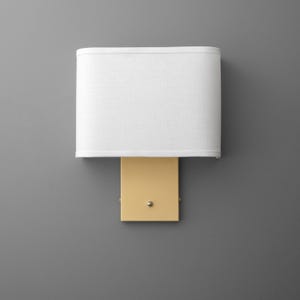 May include: A wall-mounted light fixture with a rectangular, pale yellow base and a white, oval lampshade. The fixture is mounted on a gray wall. The lampshade is made of a textured fabric.