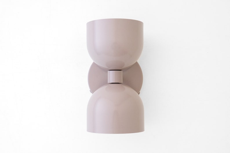May include: A modern, taupe-colored wall sconce with a unique hourglass shape. The light fixture has a rounded top and bottom, connected by a cylindrical middle section. It is mounted on a circular base.