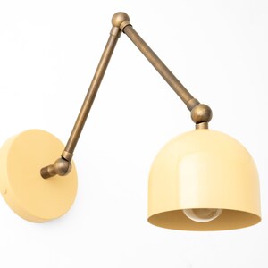 May include: Cream-colored wall-mounted lamp with a brass-toned adjustable arm and a rounded shade. The lamp has a circular base and a visible lightbulb. The design is modern and minimalist, suitable for various interior styles.