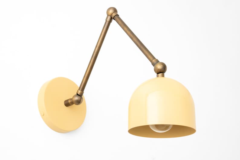 May include: Cream-colored wall-mounted lamp with a brass-toned adjustable arm and a rounded shade. The lamp has a circular base and a visible lightbulb. The design is modern and minimalist, suitable for various interior styles.