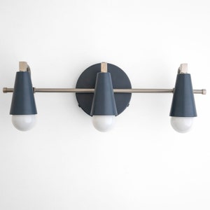 May include: A modern, three-light vanity fixture. The fixture features three navy blue cone-shaped shades with white bulbs. The fixture is mounted on a brushed nickel bar and a circular navy blue backplate. The overall design is sleek and contemporary.