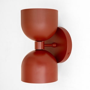 May include: A modern, rust-colored wall sconce with a rounded top and bottom, connected by a cylindrical piece. The sconce is mounted on a circular base. The fixture has a minimalist design and is suitable for indoor lighting.