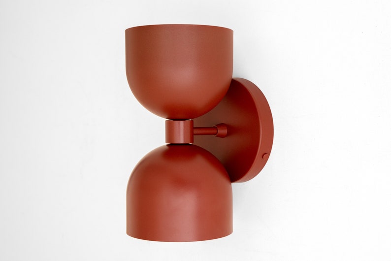 May include: A modern, rust-colored wall sconce with a rounded top and bottom, connected by a cylindrical piece. The sconce is mounted on a circular base. The fixture has a minimalist design and is suitable for indoor lighting.