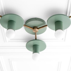 May include: A three-light ceiling fixture with a modern design. The fixture has a brass finish and green metal shades. The shades are round and have a cylindrical shape. The fixture is mounted to a white ceiling.