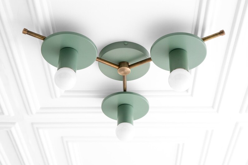 May include: A three-light ceiling fixture with a modern design. The fixture has a brass finish and green metal shades. The shades are round and have a cylindrical shape. The fixture is mounted to a white ceiling.