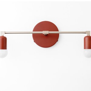 Puede incluir: Una l&aacute;mpara de pared moderna con dos bombillas. La l&aacute;mpara tiene un acabado de n&iacute;quel cepillado y una placa posterior circular roja.