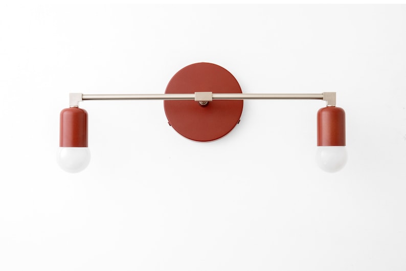 Puede incluir: Una l&aacute;mpara de pared moderna con dos bombillas. La l&aacute;mpara tiene un acabado de n&iacute;quel cepillado y una placa posterior circular roja.