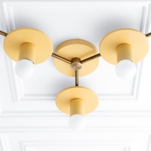 May include: A three-light ceiling fixture with a brass finish and yellow painted shades. The fixture has a modern design with three arms extending from a central hub.