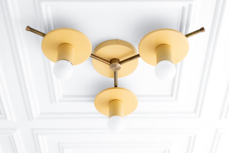 May include: A three-light ceiling fixture with a brass finish and yellow painted shades. The fixture has a modern design with three arms extending from a central hub.