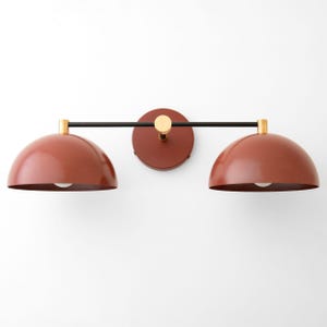 May include: A modern wall sconce with two red dome-shaped shades and a black metal bar connecting them. The sconce has a brass finish.