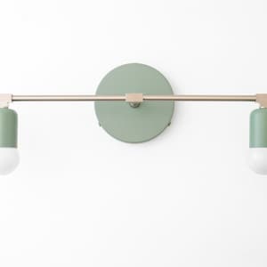 Puede incluir: Una l&aacute;mpara de pared con dos bombillas. La l&aacute;mpara tiene un acabado en n&iacute;quel cepillado y una pantalla de color verde salvia. Las bombillas son blancas.