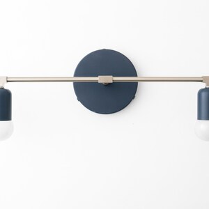 Puede incluir: Una l&aacute;mpara de pared con dos pantallas cil&iacute;ndricas azules y un brazo de metal plateado. La l&aacute;mpara tiene una placa trasera redonda y azul.