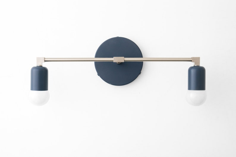 Puede incluir: Una l&aacute;mpara de pared con dos pantallas cil&iacute;ndricas azules y un brazo de metal plateado. La l&aacute;mpara tiene una placa trasera redonda y azul.