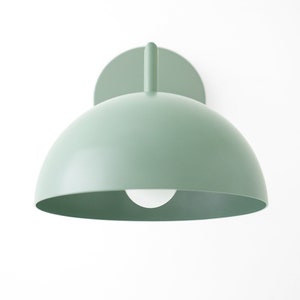 Puede incluir: Una l&aacute;mpara de pared de color verde claro en forma de c&uacute;pula con una bombilla blanca. La l&aacute;mpara tiene un dise&ntilde;o simple y moderno.