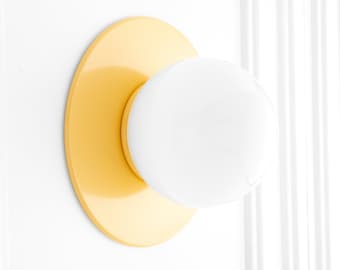 May include: A wall-mounted light fixture featuring a circular, smooth yellow base plate and a spherical, opaque white glass globe shade. The design is simple and geometric, with the white globe centered on the flat, bright yellow circular mount.