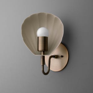 May include: A wall-mounted light fixture with a seashell-shaped shade in a cream color. The fixture has a brass-colored base and a curved arm holding a white light bulb. The fixture is designed to be mounted on a wall.