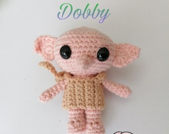 May include: A light pink crocheted doll with large black eyes, oversized ears, and a tan tunic. The word Dobby is printed in a gradient font above the doll, and a small cartoon illustration of the character is positioned in the bottom right corner.