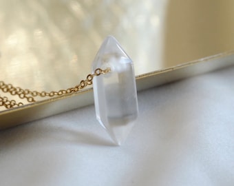 Puede incluir: Un collar con colgante de cristal de cuarzo transparente con una cadena dorada. El cristal es un punto de doble terminación y la cadena es delicada. El collar se muestra sobre una superficie clara.