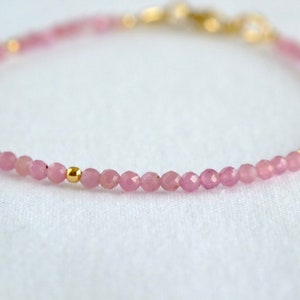 May include: A delicate bracelet featuring small, faceted pink gemstones and gold-coloured beads. The bracelet is strung together with gold-coloured beads interspersed throughout. The clasp is also gold-coloured. The bracelet is displayed on a white surface.