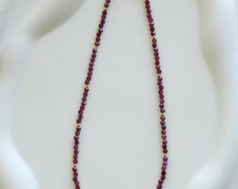May include: A delicate necklace featuring small, faceted, deep red beads strung together. The necklace has gold-colored accents and a gold-colored chain and clasp. The jewelry is displayed on a white surface.