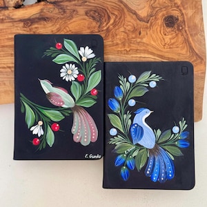May include: Two black notebooks with hand-painted bird designs. One features a pink and green bird with white flowers and red berries. The other has a blue bird with blue flowers and berries. Both are signed by E. Gunko.