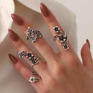 Puede incluir: Una mano con cuatro anillos plateados con diseños florales. Los anillos presentan intrincados detalles de flores y hojas, con un anillo que se extiende por el dedo. Los anillos se presentan sobre un fondo neutro.