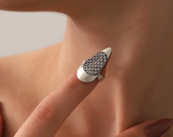 May include: A silver-toned finger ring with a white, elongated oval top. The top is adorned with a pattern of small, dark stones. The ring is worn on a finger, showcasing its design and craftsmanship. The ring's design is elegant and modern.