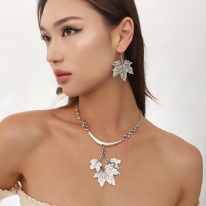 Puede incluir: Conjunto de collar y pendientes de tono plateado con diseño de hoja. El collar tiene una barra curva con tres hojas colgando. Los pendientes son pendientes de gota simples con un diseño de hoja única.