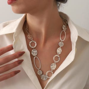 May include: Silver-toned necklace featuring a series of textured, circular, and oval links. The necklace is worn against a white collared shirt. The links have a hammered appearance, creating a unique, eye-catching design.