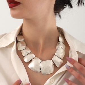 Puede incluir: Un collar plateado con colgantes cuadrados grandes. El collar se lleva sobre una camisa blanca con cuello. Los colgantes tienen una superficie texturizada y están conectados por pequeños eslabones.