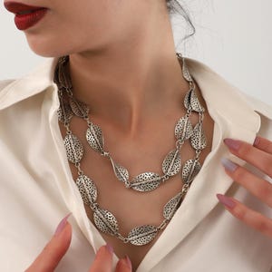 Könnte beinhalten: Eine silberne mehrreihige Halskette mit ovalen Gliedern, die ein strukturiertes Blattdesign aufweisen.