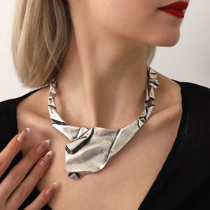 May include: A silver metal necklace with a unique, abstract design. The necklace is made of a single piece of metal that is folded and twisted into a shape that resembles a crumpled piece of paper. The necklace is a statement piece that would add a touch of modern style to any outfit.