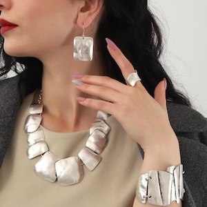 Peut inclure: Un ensemble de bijoux en argent composé d'un collier, d'un bracelet et de boucles d'oreilles imposants. Les pièces sont en métal texturé et présentent un design géométrique et moderne.