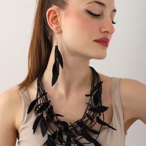 May include: A black leather necklace with multiple layers of leaves. The necklace is worn by a person with long dark hair and black dangle earrings.