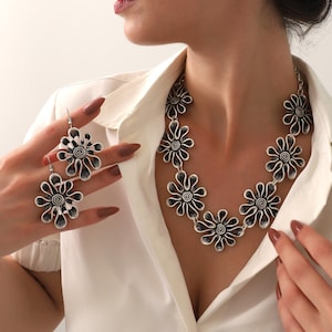 Op de afbeelding: Een zilverkleurige bloemen sieradenset, bestaande uit een ketting en oorbellen. De ketting heeft meerdere bloemvormige hangers en de oorbellen hebben een vergelijkbaar ontwerp. De sieraden worden gedragen bij een wit overhemd.