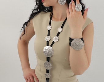 May include: A silver necklace with large, textured, round pendants hangs from a black cord. The necklace is paired with matching earrings and a bracelet.