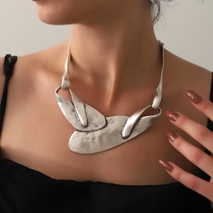 May include: A silver-toned statement necklace with an abstract design. The necklace features a textured, hammered finish and a bold, asymmetrical shape. The necklace is worn against a black top.