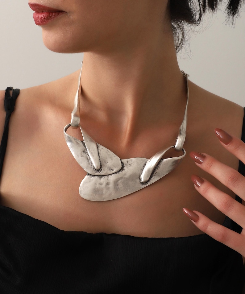 May include: A silver-toned statement necklace with an abstract design. The necklace features a textured, hammered finish and a bold, asymmetrical shape. The necklace is worn against a black top.