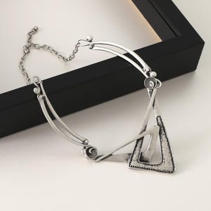 May include: A silver-toned necklace with a geometric triangle pendant. The pendant is made of two overlapping triangles with a textured surface.